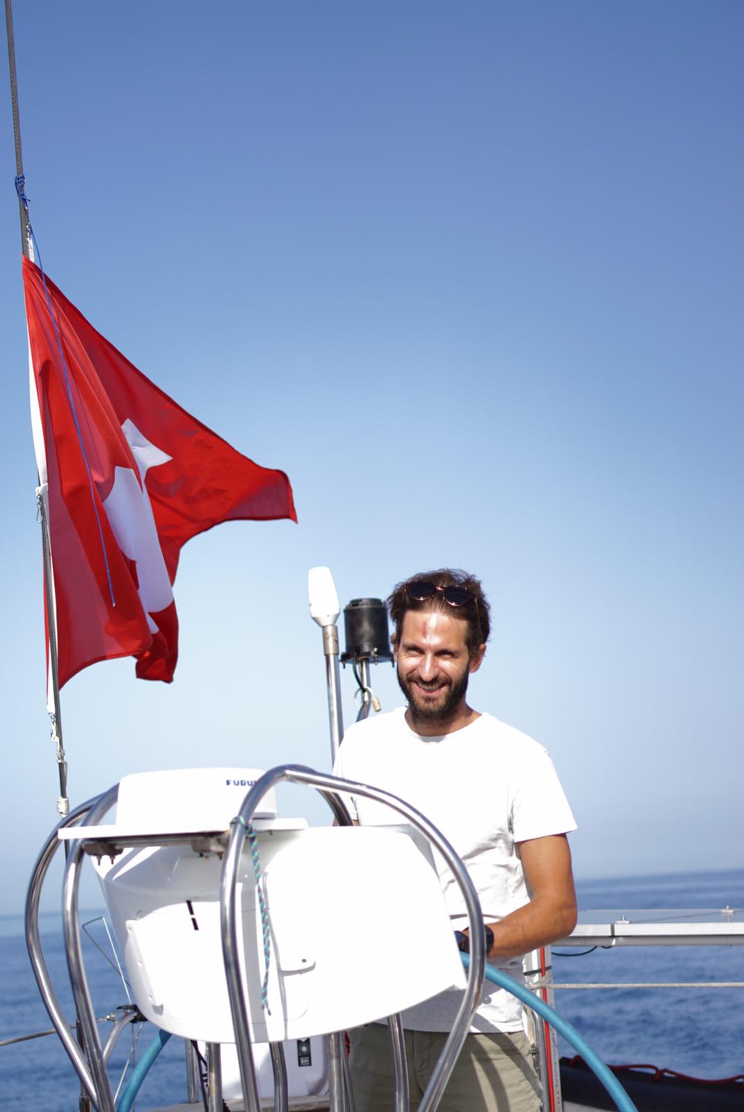 A smiling sailor at the helm of a sailboat under a clear blue sky, a red flag flying beside him